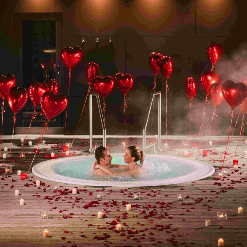 Couple Dans Le Jacuzzi Extérieur Du Spa Privatif Avec La Décoration Romantique, Pétales De Roses, Bougies Autour, Ballons En Forme De Coeur